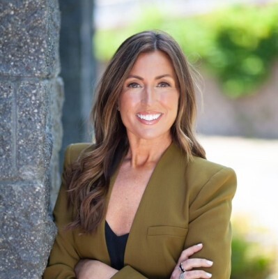 Woman with long brown hair in an olive blazer smiles outdoors, leaning against a stone wall.