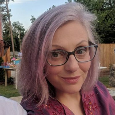 Woman with light purple hair, glasses, and a slight smile, outdoors with trees and a wooden fence in background.
