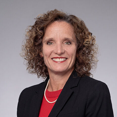 Woman with curly brown hair in a black blazer and red shirt, smiling at the camera against a gray background—an accomplished Interim CFO in San Francisco with extensive experience in both non-profit and for-profit corporate finance.