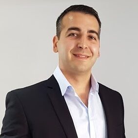 Man in a dark suit jacket and white shirt smiling in front of a plain light background, exemplifying the professionalism of an experienced Interim CFO with expertise in both non profit and for-profit corporate finance in San Francisco.