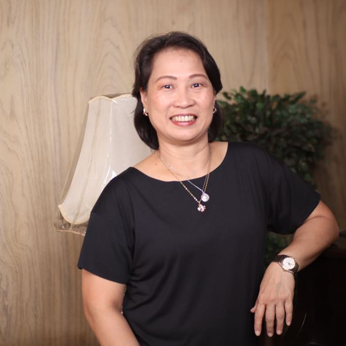 A woman in a black shirt, who serves as an Interim CFO, smiles while standing indoors near a lamp and a green plant.