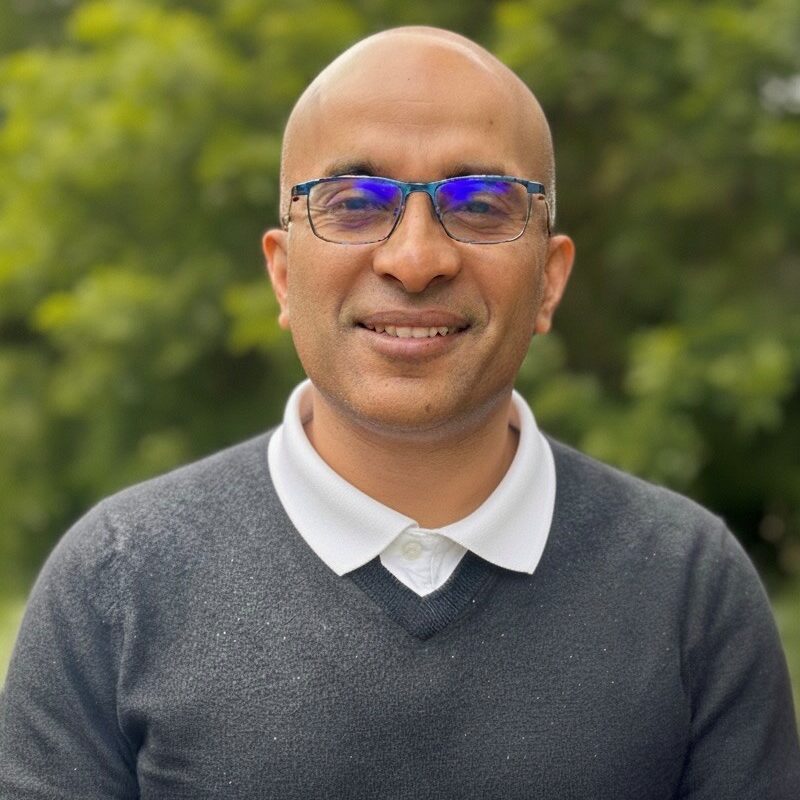 A smiling man with glasses and a bald head, wearing a dark sweater over a white collared shirt, outdoors.