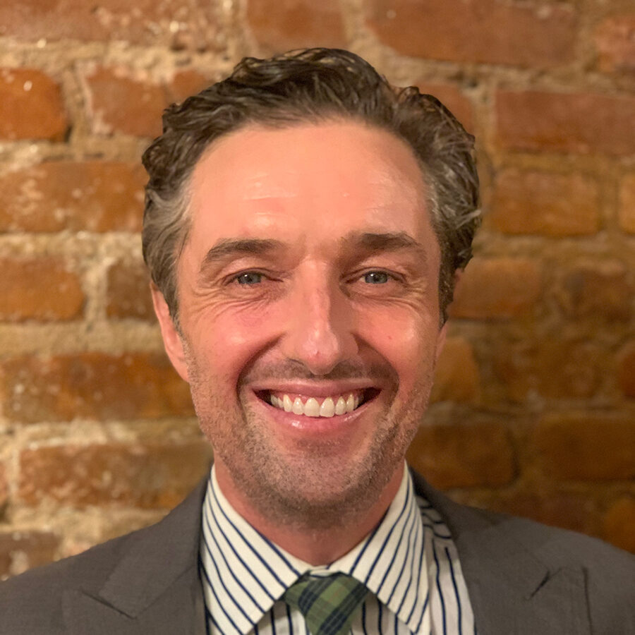 Smiling man in a suit and striped shirt standing in front of a brick wall.