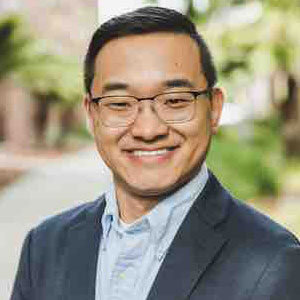 Smiling man with glasses wearing a suit jacket, outdoors with greenery in the background; an experienced Interim CFO in San Francisco, skilled in both non profit and for-profit corporate finance.