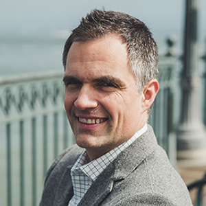 Man in a gray blazer smiling outdoors by a metal fence with water in the background.
